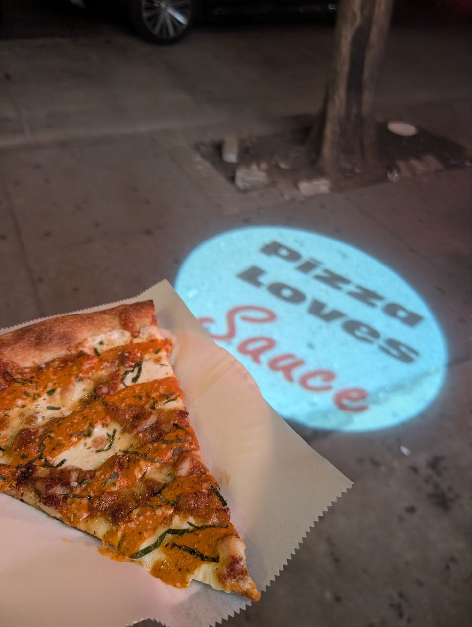 Slice of chicken vodka pizza on wax paper with the Pizza Loves Sauce logo projected on the sidewalk in New York City at night